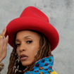 Sa-Roc wearing a red wide-brim hat and multicolored patterned jacket, posing against a gray backdrop with long locs and gold hoop earrings.