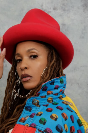 Sa-Roc wearing a red wide-brim hat and multicolored patterned jacket, posing against a gray backdrop with long locs and gold hoop earrings.
