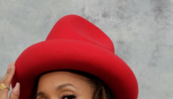 Sa-Roc wearing a red wide-brim hat and multicolored patterned jacket, posing against a gray backdrop with long locs and gold hoop earrings.