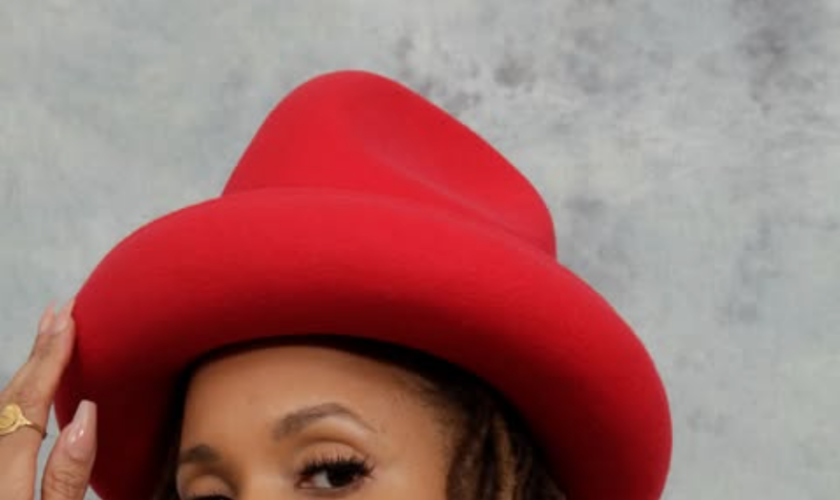 Sa-Roc wearing a red wide-brim hat and multicolored patterned jacket, posing against a gray backdrop with long locs and gold hoop earrings.