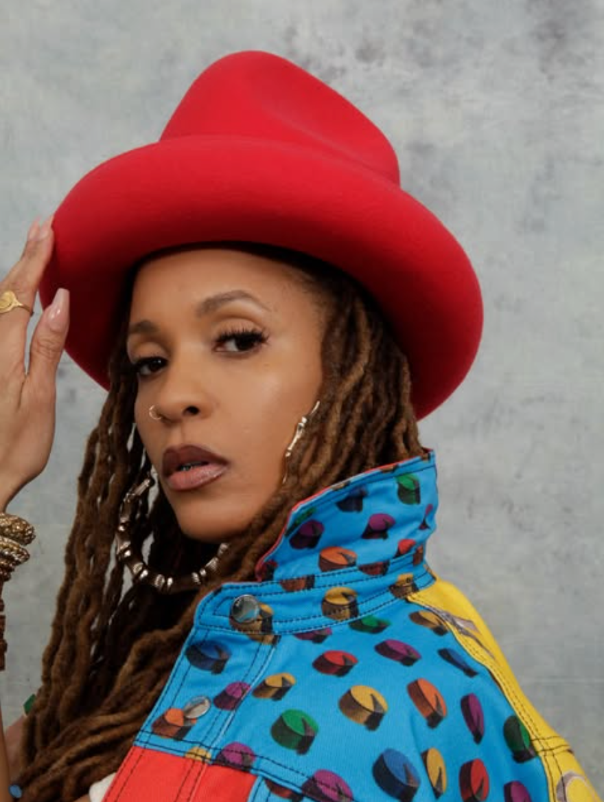 Sa-Roc wearing a red wide-brim hat and multicolored patterned jacket, posing against a gray backdrop with long locs and gold hoop earrings.